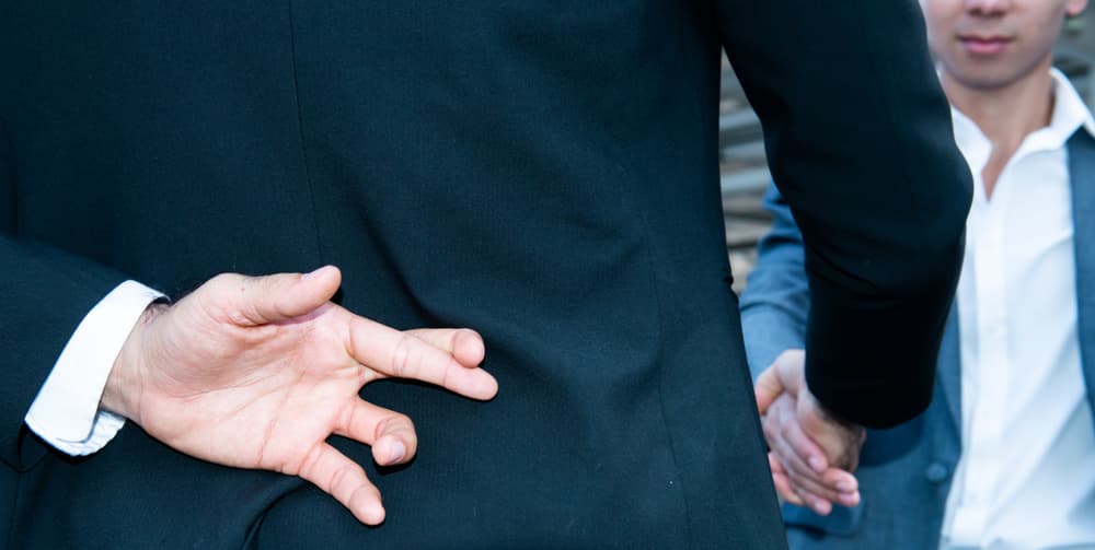 Lawyer crossing fingers behind back during handshake, representing dishonest settlement tactics by insurance attorneys.