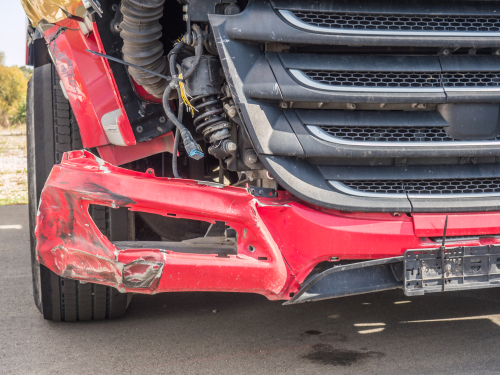 Close-up of a red commercial truck with a broken front bumper and visible damage after a collision, parked on a paved surface.