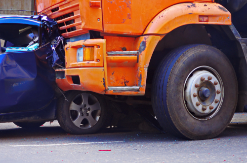 Orange commercial truck crushing the front of a blue car in a severe collision, showing major front-end damage and a trapped wheel.