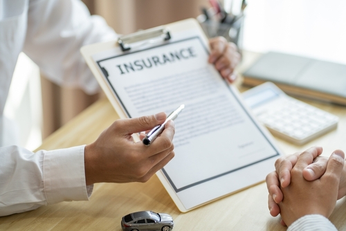 A person holding a clipboard with an insurance document, explaining details to another person across a desk with a toy car and calculator present.