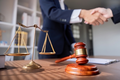 Lawyer and client shaking hands across a desk with a gavel and scales of justice in the foreground.