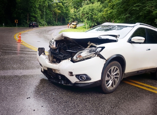 A white SUV with a severely crumpled front end sits on a wet, winding road after a crash, with warning cones and another vehicle visible in the distance.
