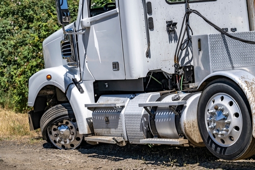 White semi-truck parked on the roadside with a blown front tire, showing visible damage and dirt.