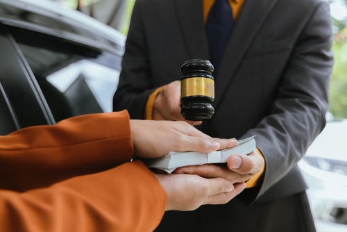 Person handing a stack of money to another person holding a judge’s gavel near a car, symbolizing a legal settlement or payment.