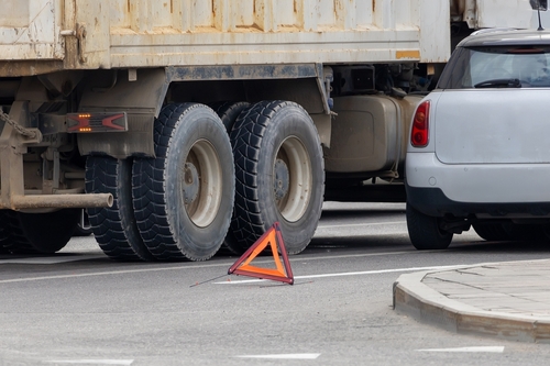 Close view of a parked truck and a car on a city street with a reflective warning triangle placed on the road behind the truck’s rear wheels.
