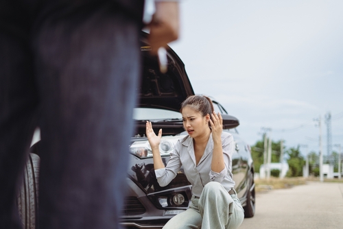 A frustrated young woman stands beside her car with the hood open on the roadside, gesturing with both hands as she speaks to a person approaching.