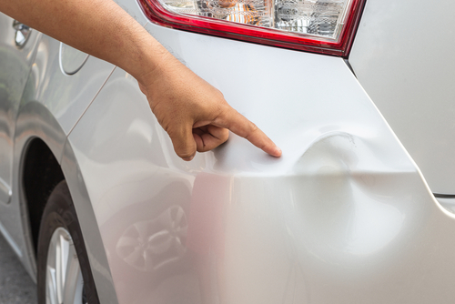 Hand pointing to a small dent on the rear bumper of a silver car.
