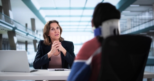 A woman in a black blazer sitting at a table with a laptop, looking attentively at a person wearing a headset and neck support, who is facing away from the camera in a modern office setting.