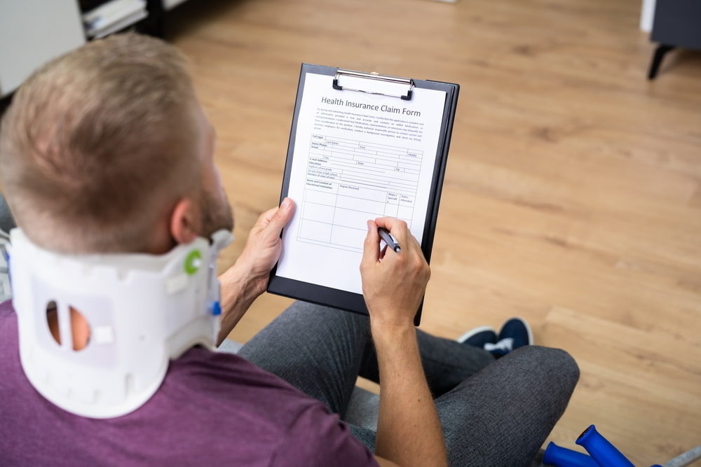 Man wearing a neck brace filling out a health insurance claim form after suffering injuries in an accident