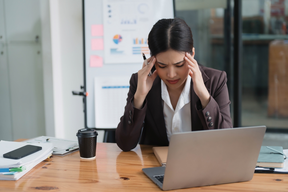Office worker holding her head while experiencing a headache at her desk in the workplace.