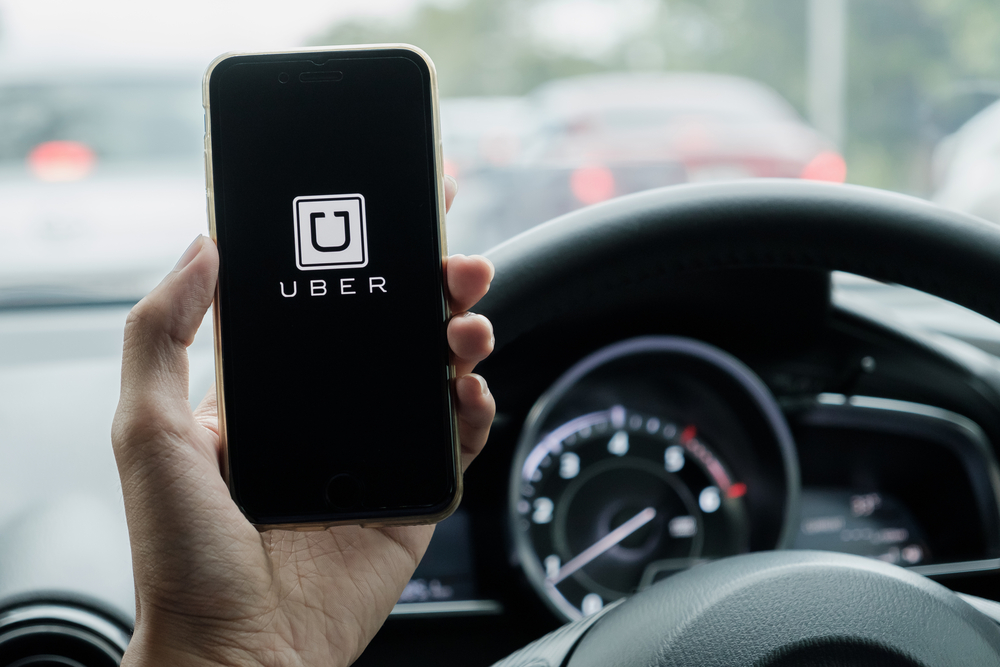 A uber driver hand holding Uber app showing on iPhone 6s on road and black car, Uber is smartphone app-based transportation network.