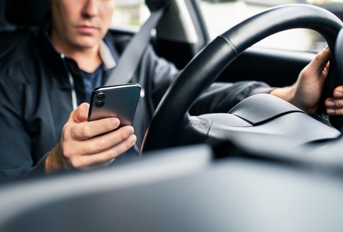 A man holding a smartphone in his right hand while driving a car with his left hand on the steering wheel.