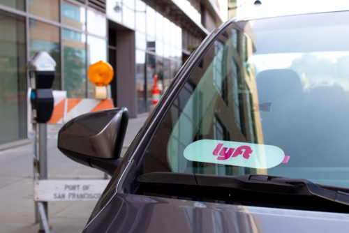 A gray car with a Lyft logo sticker displayed on the windshield, parked near a construction zone with caution signs and barriers in the background.