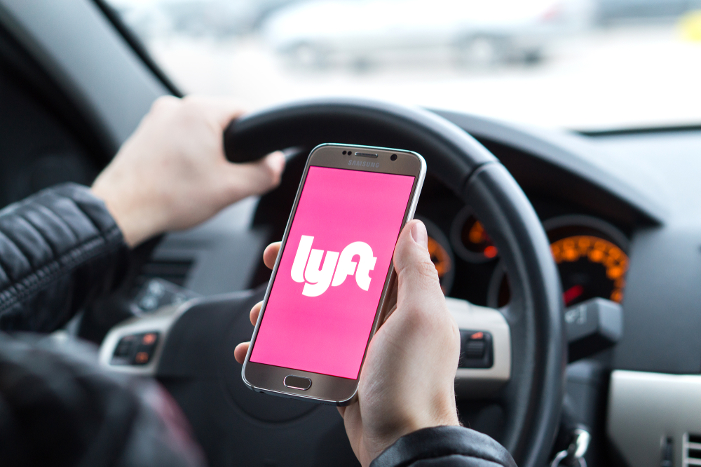 Rideshare driver holding a smartphone with the Lyft app while seated behind the steering wheel
