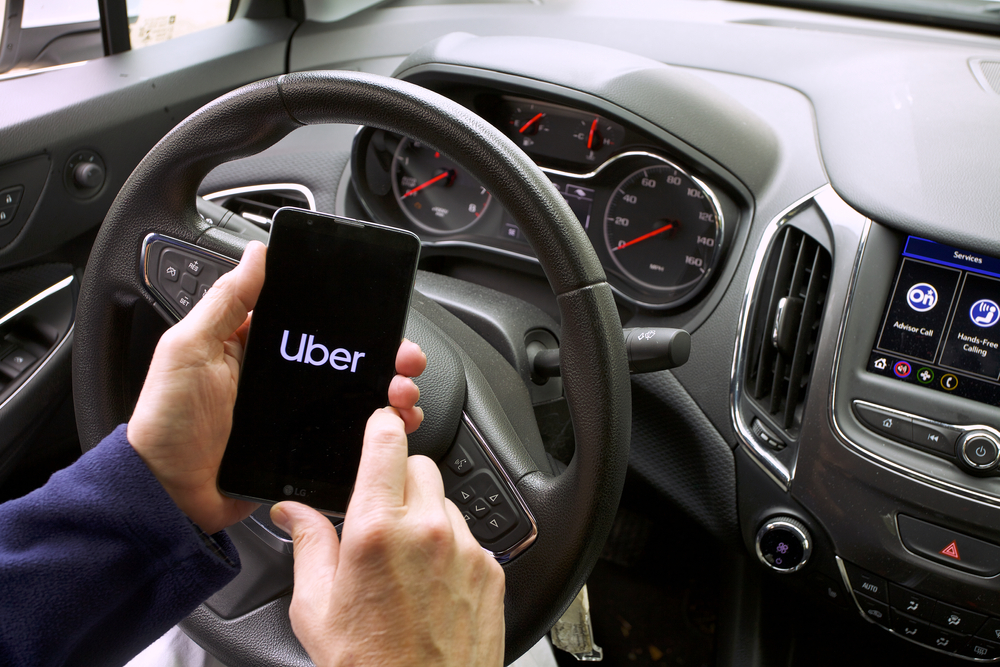 Driver holding a smartphone displaying the Uber app while inside a parked vehicle