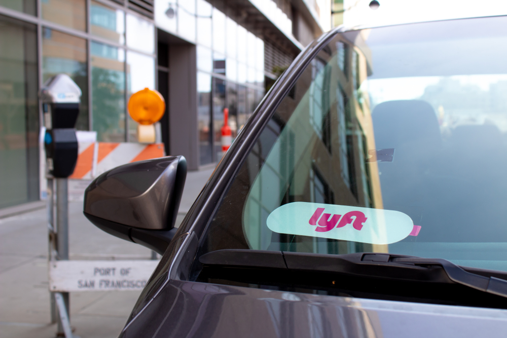 Close-up of a car's side mirror with a pink and white Lyft decal displayed on the windshield. Background shows a city street with construction barriers.