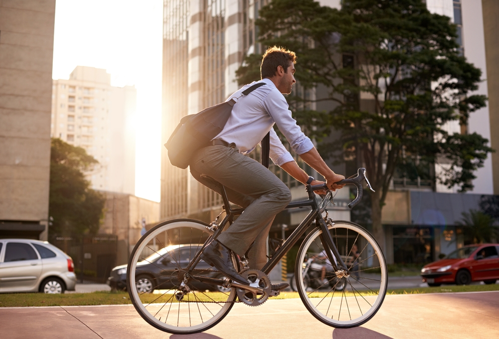 A man in a light blue shirt and gray pants riding a bicycle on a city street during sunset, with buildings and cars visible in the background.
