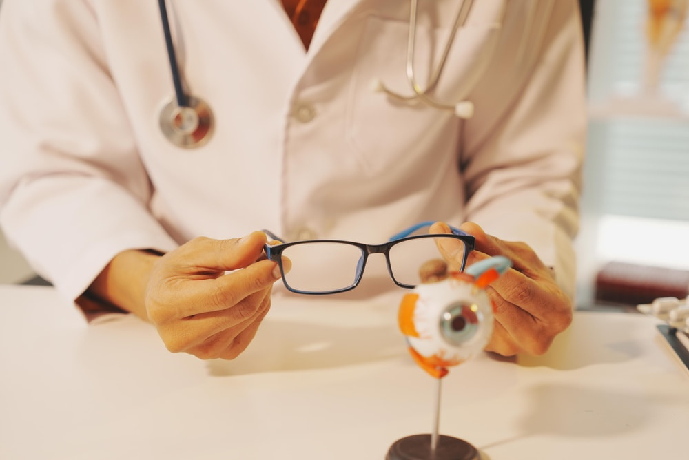 A person in a white lab coat holding blue eyeglasses in one hand and an eye model on a stand in the other hand, positioned on a white table