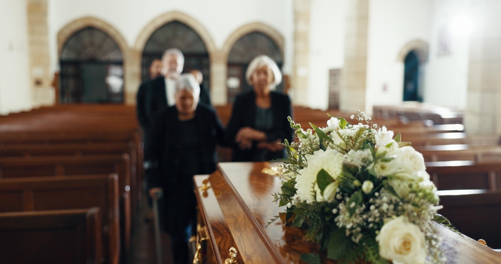 A casket adorned with a floral arrangement in a church, with three people walking in the background.