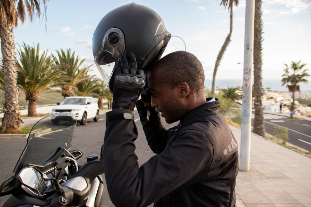 A man wearing a black jacket and gloves holding a motorcycle helmet near a motorcycle on a palm tree-lined street.