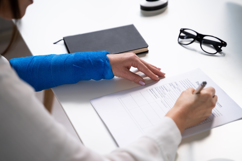 A person with a blue cast on their left arm writing on a document while holding a pen. A black notebook and glasses are placed nearby on the white table.