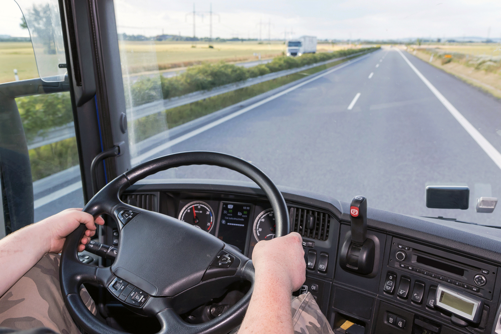 Hands gripping a large steering wheel inside a truck cabin, showing the dashboard and an open highway ahead.
