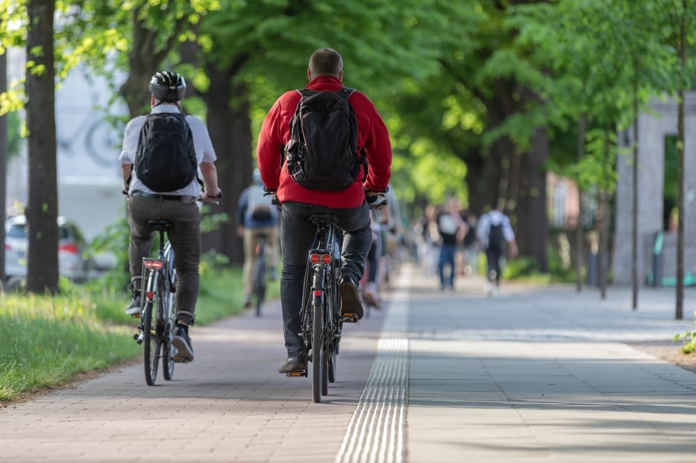 Two adults riding bicycles on a tree-lined path, one without a helmet.